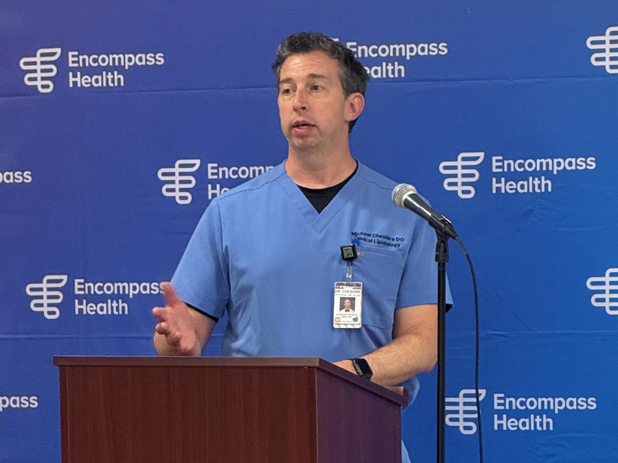 A man in blue scrubs speaks at a podium with a microphone, set against a blue Encompass Health backdrop. His expression is focused and professional.