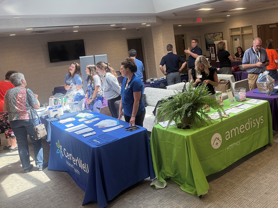A health fair with tables and colorful banners displaying CarerWell Home Health and Amedisys Hospice. Participants engage, with a lively, collaborative atmosphere.