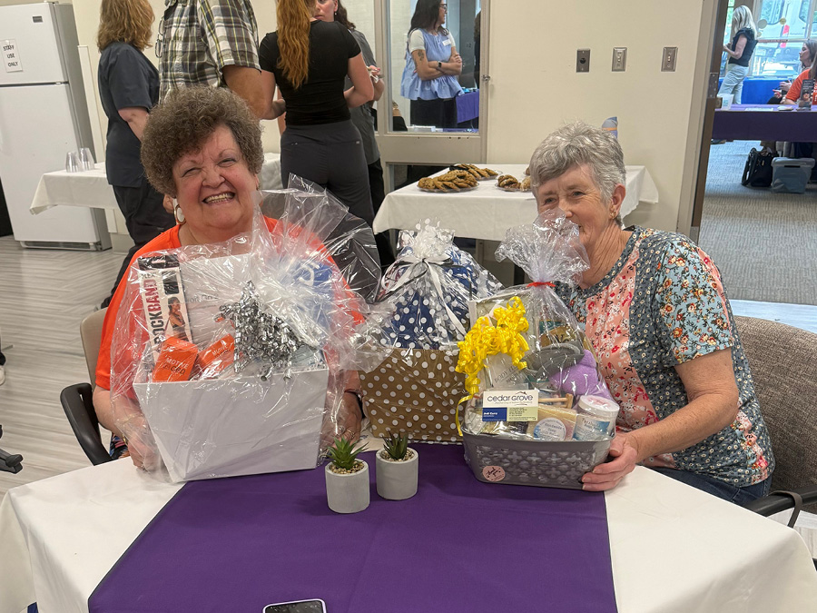 Two elderly women are seated at a table, smiling and holding gift baskets with decorative wrapping. The room has people mingling in the background and a table with snacks. The atmosphere is cheerful and festive.