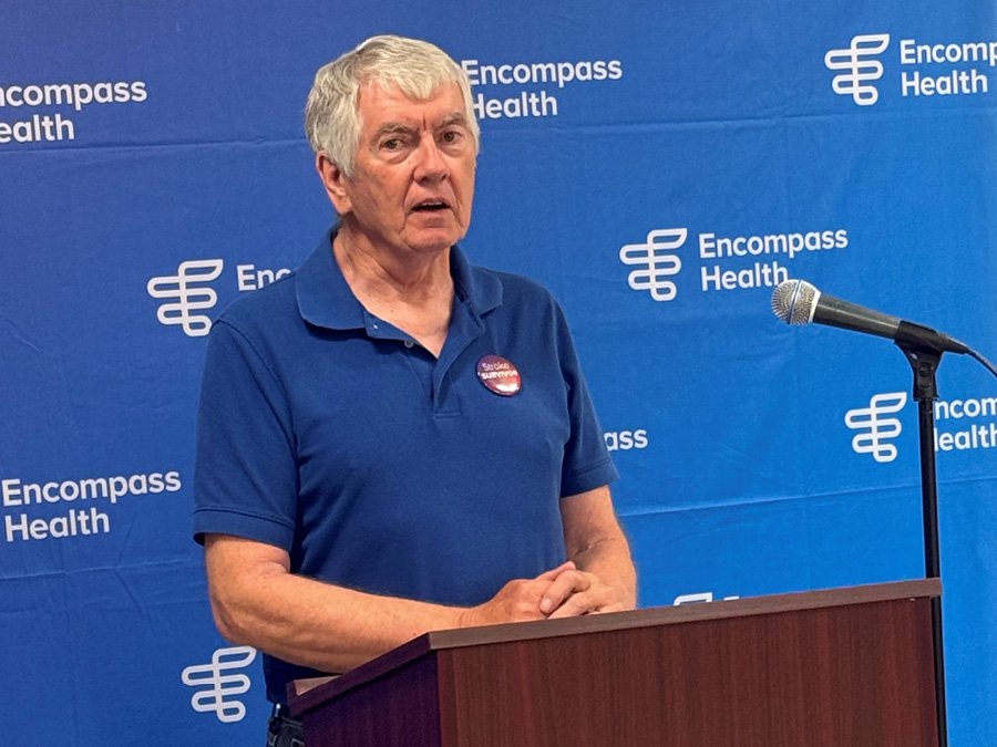 A person with gray hair speaks at a podium with a microphone. They wear a blue shirt with a Stroke Prevention button. The backdrop features the Encompass Health logo.