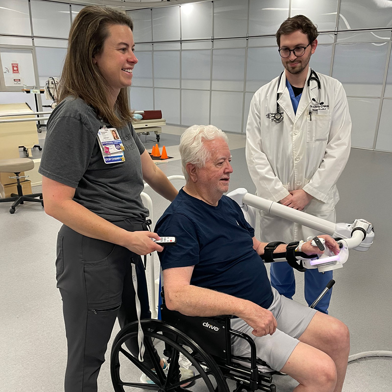 A healthcare professional assists a smiling elderly man in a wheelchair using a medical device, while a doctor observes, conveying a supportive atmosphere.