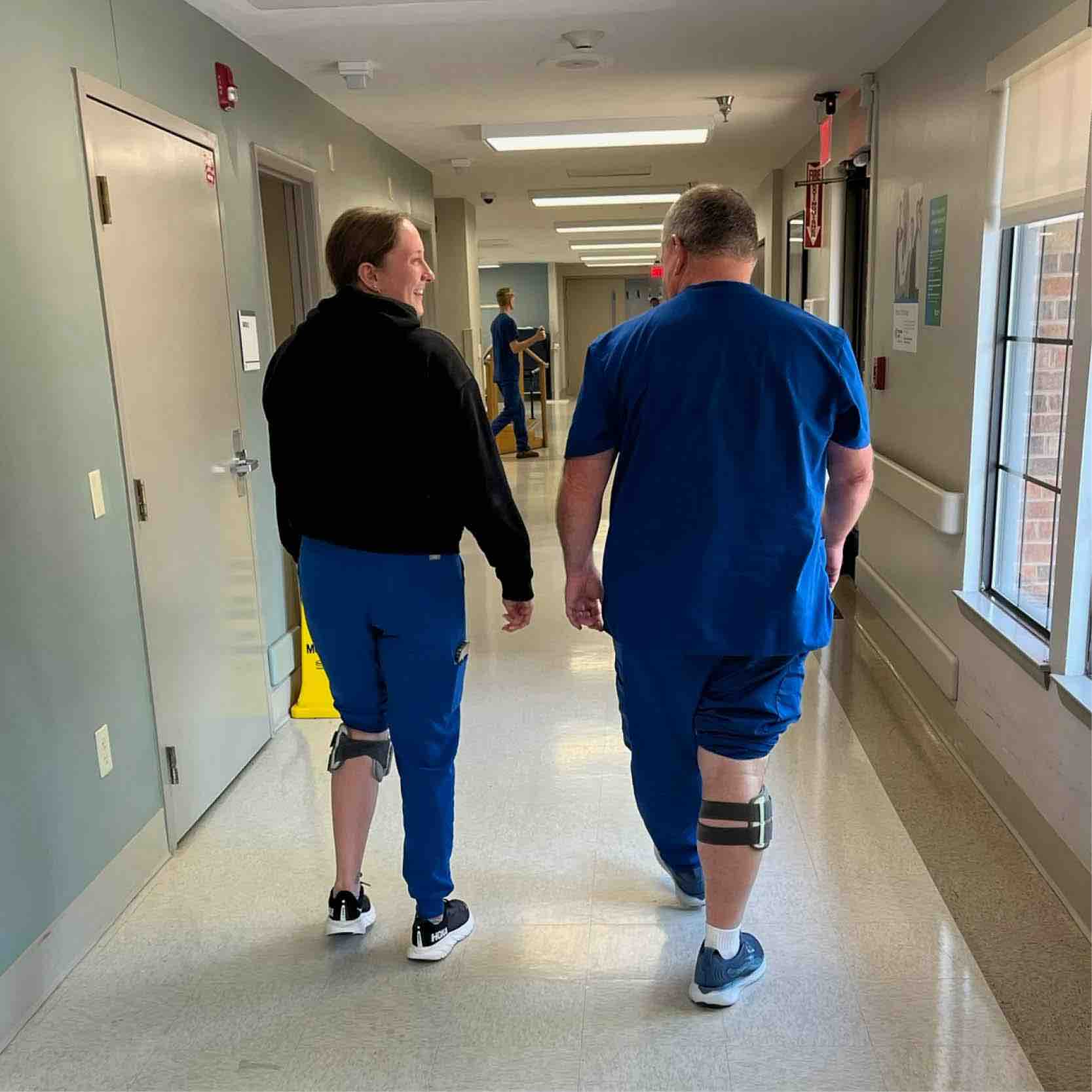 Patient and clinician walk in rehabilitation hallway using Bioness L300 system on lower leg, supporting functional electrical stimulation to improve gait and mobility after stroke
