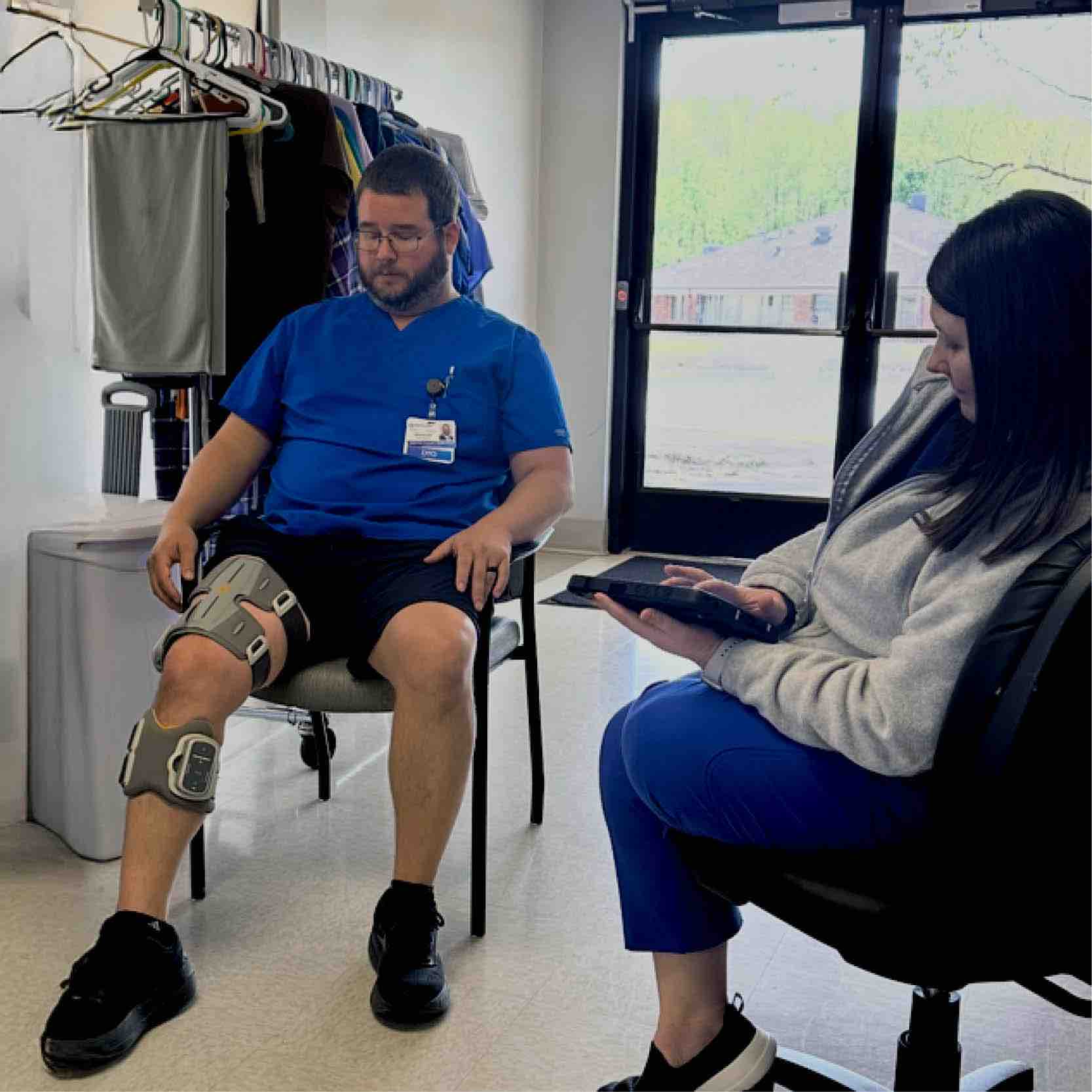 Clinician demonstrates Bioness L300 device on patient seated in therapy room, using tablet to adjust stimulation settings and support personalized neurorehabilitation and functional mobility