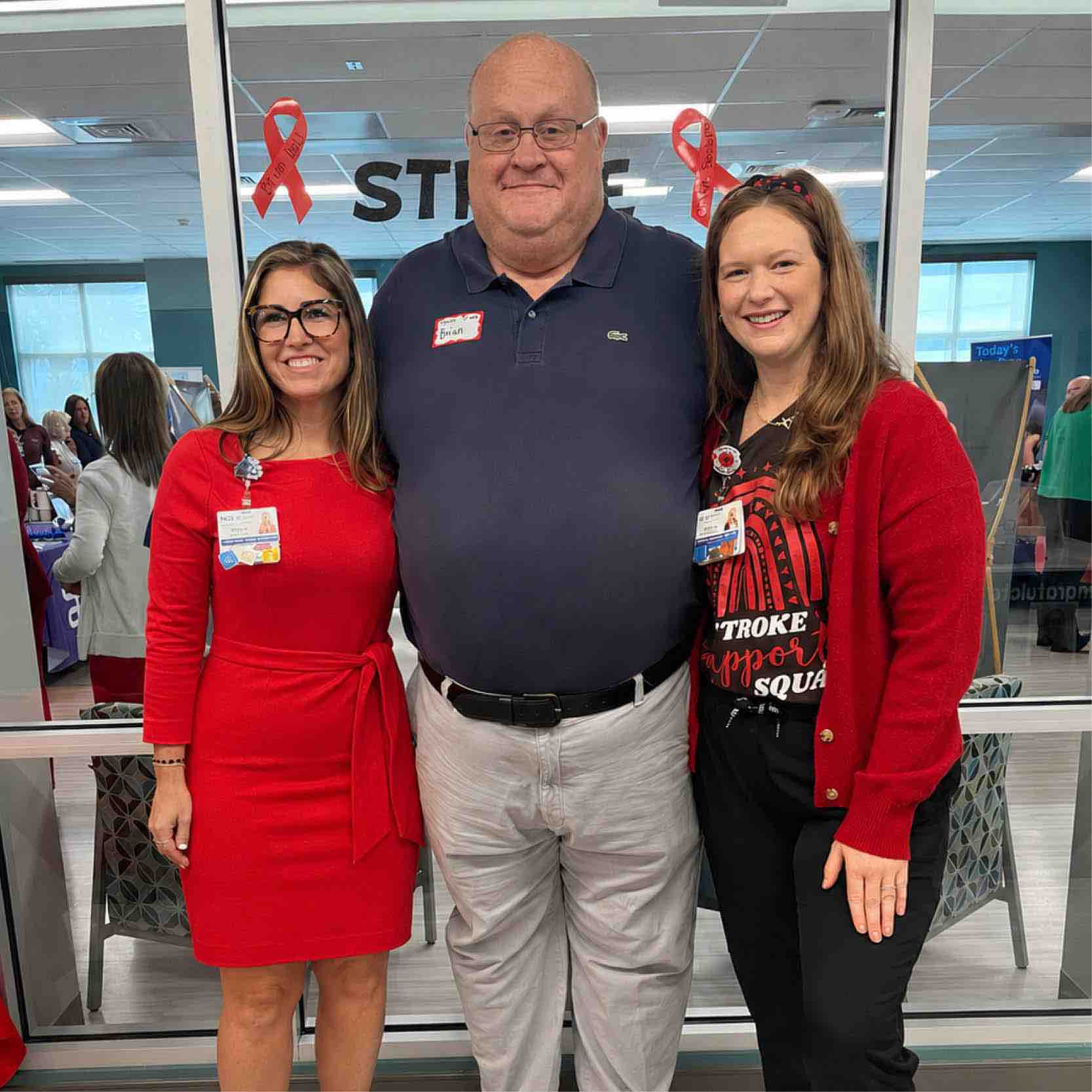 Three people stand together at a healthcare event with red stroke awareness ribbons in the background, highlighting community involvement in stroke rehabilitation and support services.