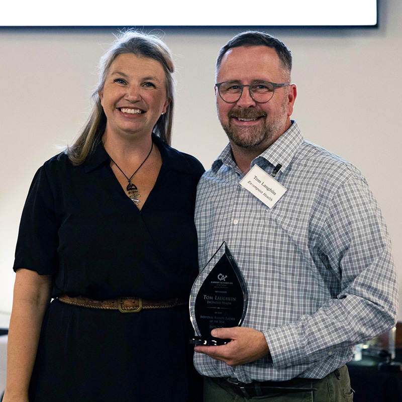 Two people stand together smiling; the man on the right holds a glass award and wears a name tag, while the woman on the left stands with her arm around him.