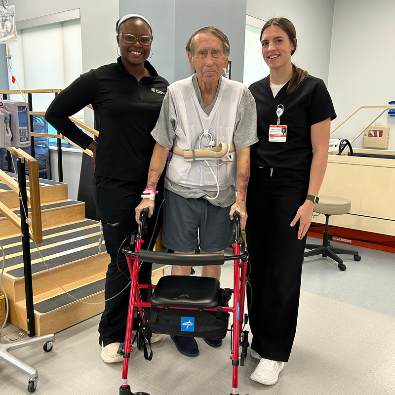 Older adult using a red walker in a rehabilitation gym, standing with two healthcare team members beside a therapy staircase.
