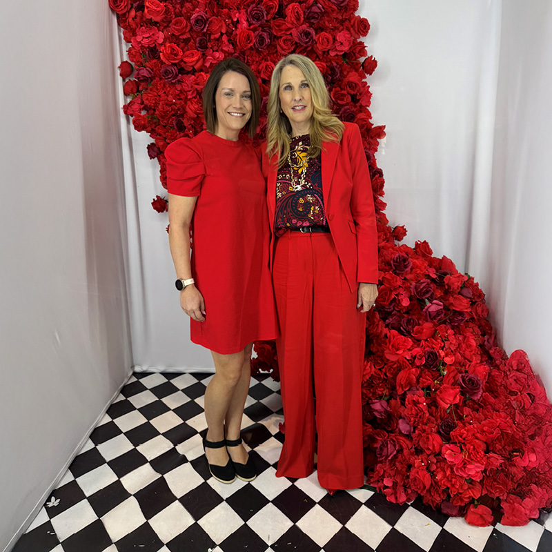 Two women in red outfits stand on a black and white checkered floor, backed by a cascade of vibrant red roses, creating an elegant, festive atmosphere.