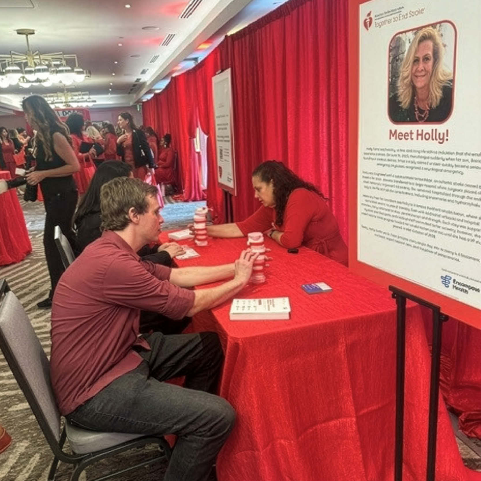 Palm Beach Go Red event booth with participant engaging in a hands-on activity at a red-draped table, surrounded by attendees learning about heart health and stroke awareness in a community setting.