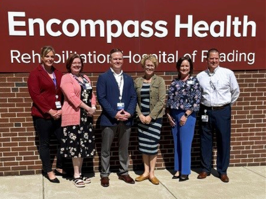 Six people pose in front of the Encompass Health Rehabilitation Hospital of Reading sign outside a brick building.