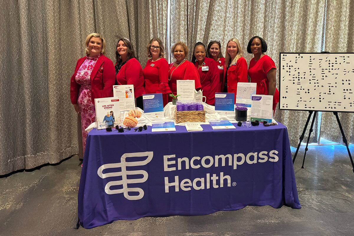 A group of eight women stands behind a table with a purple Encompass Health cloth, displaying health information and materials. All are wearing red, conveying unity and support. The setup is professional, with a large board and curtain backdrop, suggesting an event or presentation.