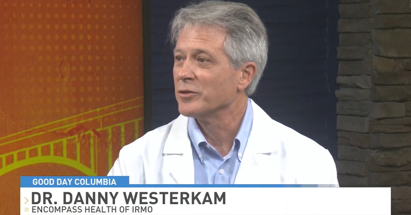 Man with gray hair wearing a white lab coat, speaking on a TV show. Background features an orange and brick texture. Text overlay shows the show name and a name.