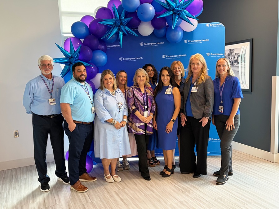 A group of ten people, wearing business attire in shades of blue, stand smiling in front of a blue Encompass Health backdrop with purple and blue balloons.