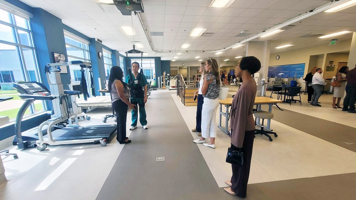 A spacious, well-lit rehabilitation center with large windows. Several people engage in conversation, surrounded by medical equipment and exercise machines. The atmosphere is professional and collaborative.