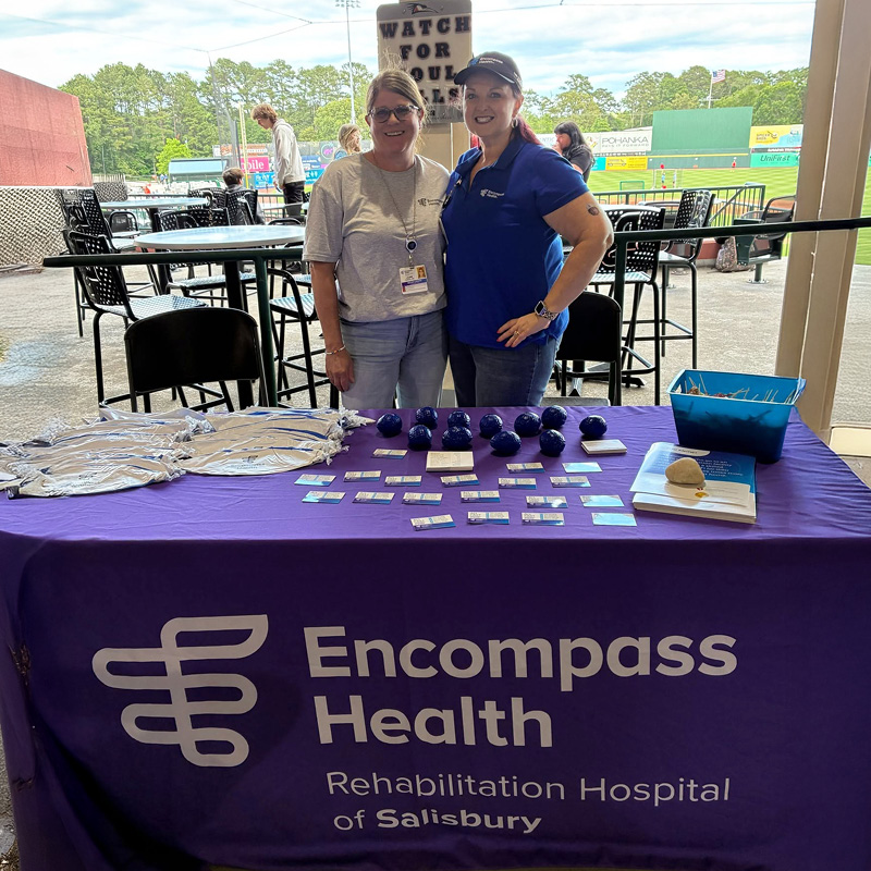 Two smiling women stand behind a display table with Encompass Health Rehabilitation Hospital of Salisbury banner, showcasing promotional items.