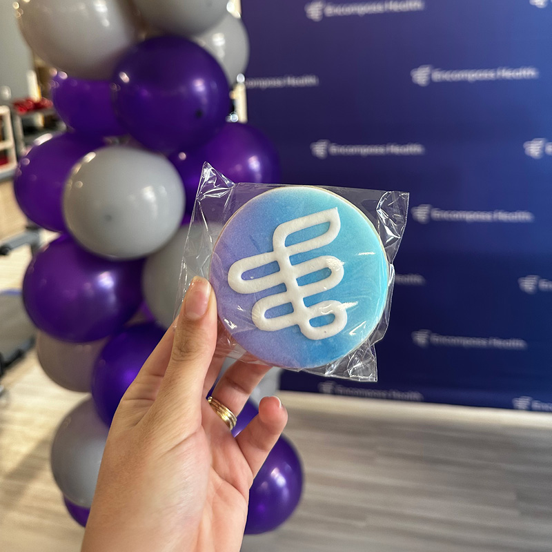 A hand holds a frosted blue cookie with an Encompass Health design, in front of a purple and gray balloon column. Background features a branded backdrop.