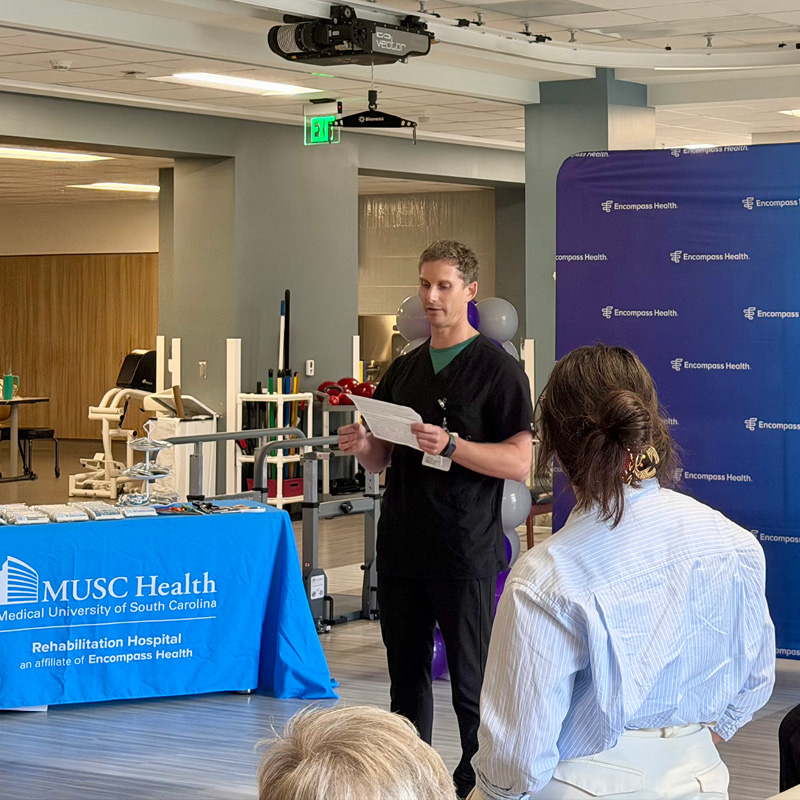 A man in black scrubs speaks at an event in a hospital setting. A table with a blue cloth reads MUSC Health. Attendees listen attentively.