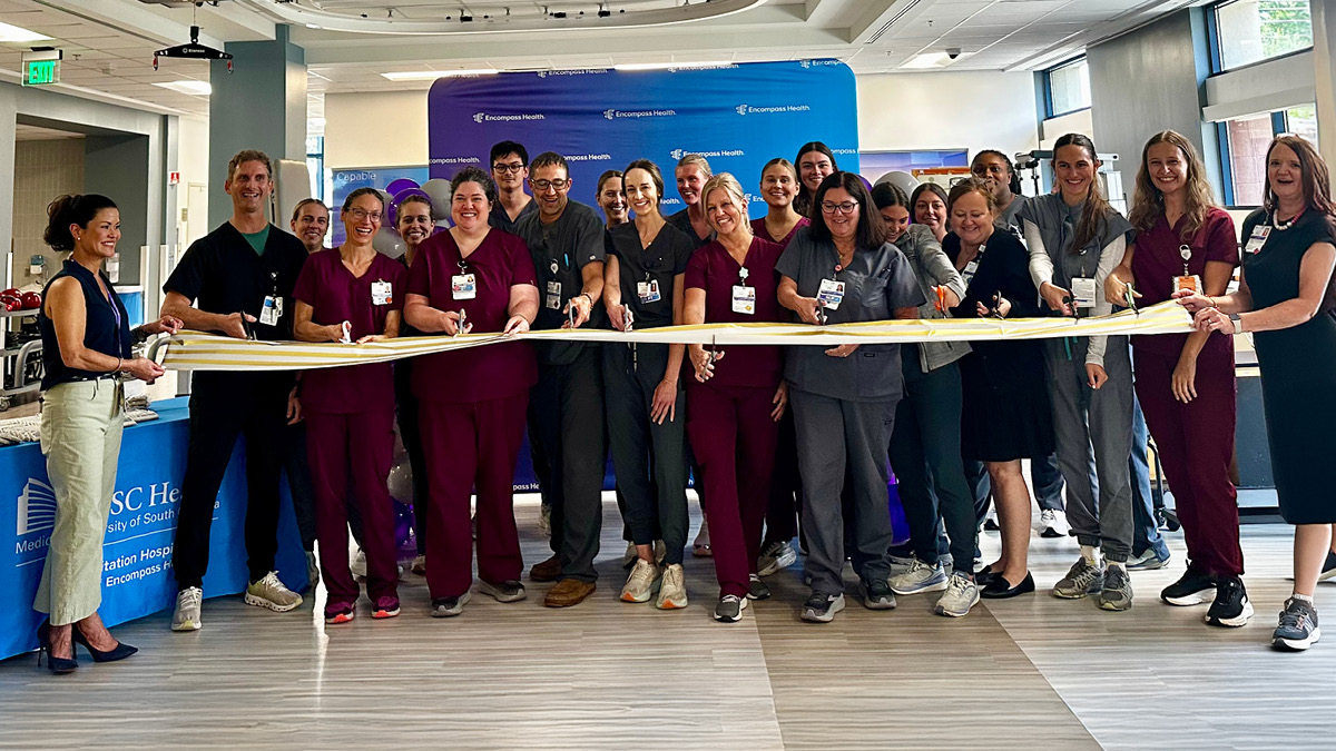 A group of smiling healthcare professionals in uniforms participate in a ribbon-cutting ceremony indoors. The atmosphere is celebratory and joyful.