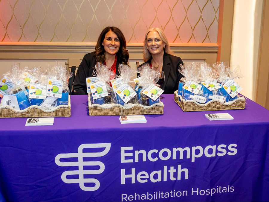 Two women smiling behind a table covered with a purple Encompass Health cloth, showcasing gift baskets with hygiene items. The setting is professional and welcoming.