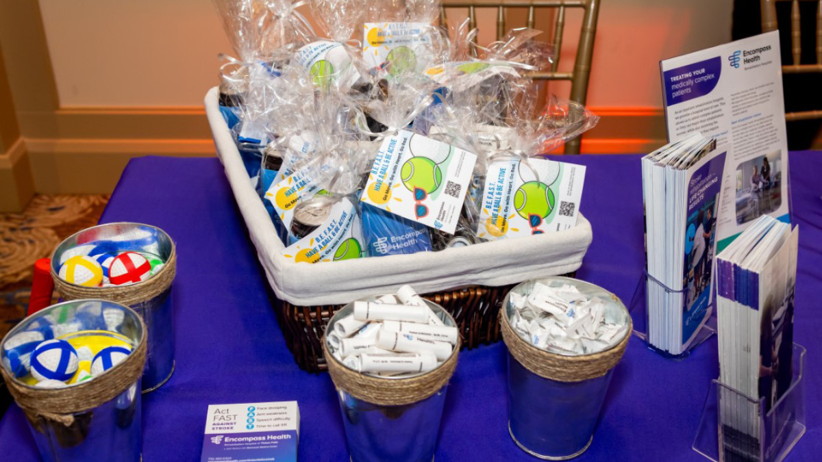 A display table with a purple cloth holds a basket of wrapped promotional items, metal buckets with stress balls, and brochures for Encompass Health.