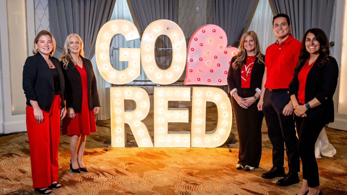 Five people stand smiling next to a large illuminated Go Red sign with a heart. They are wearing red and black, conveying a celebratory tone.