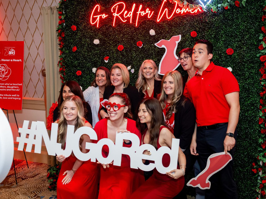 Group of smiling people in red clothing pose holding a large #NJGoRed sign. Background features greenery with red roses and a neon Go Red for Women sign.