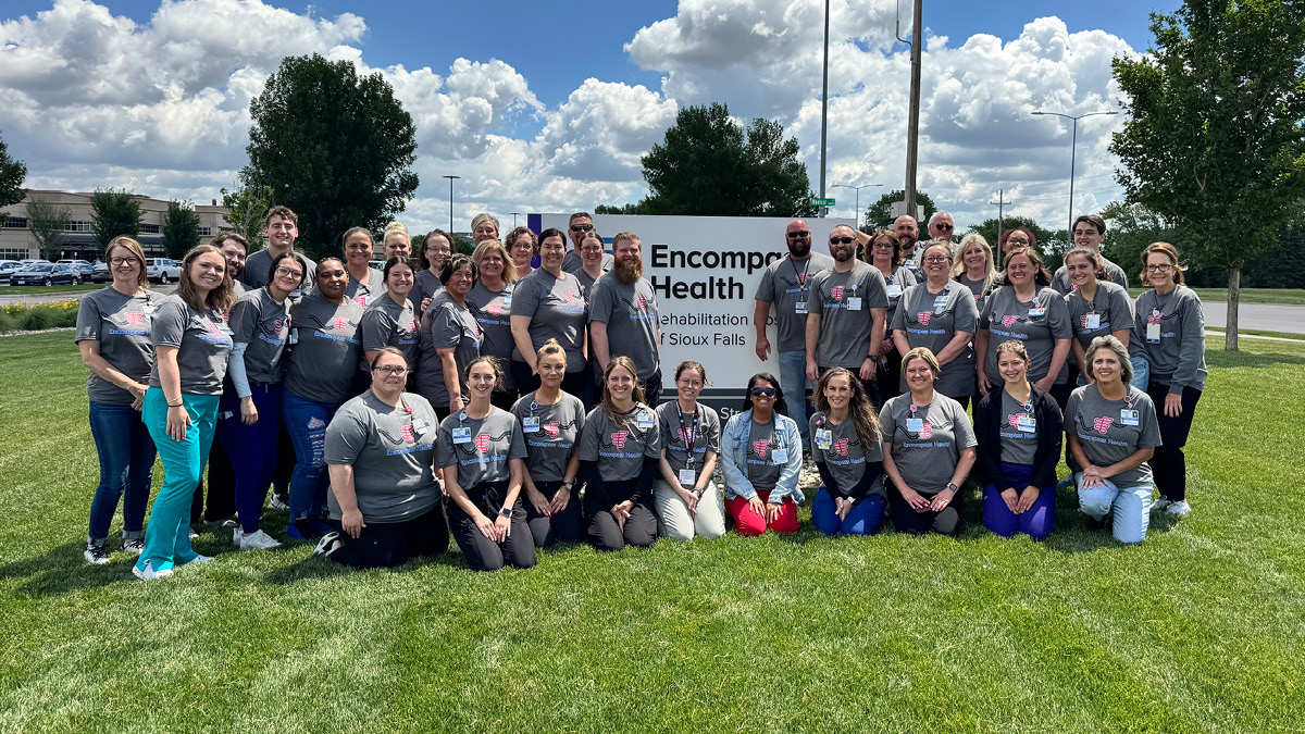 A large group of healthcare team members wearing matching gray shirts pose outdoors on a grassy lawn beside an Encompass Health Rehabilitation Hospital of Sioux Falls sign.