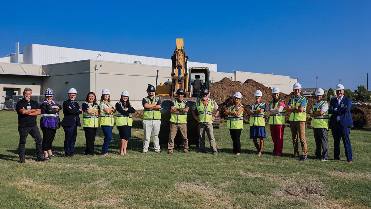 Construction and healthcare team members in hard hats stand at a construction site with an excavator and building in the background.