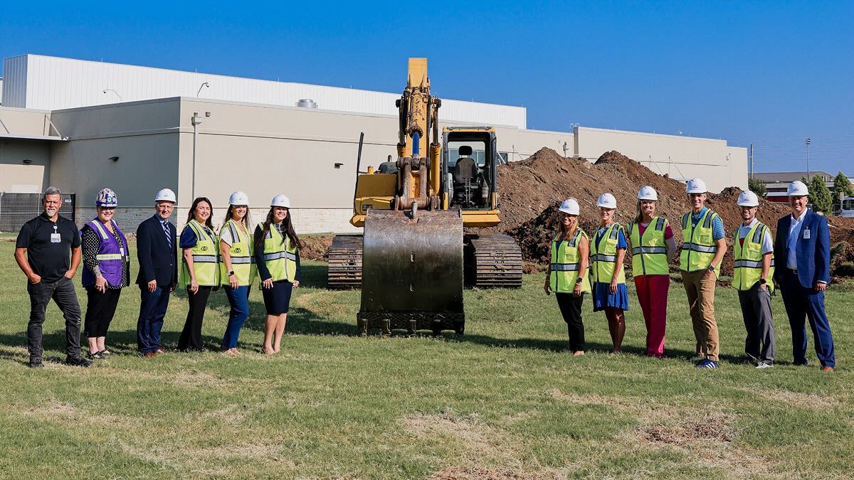 People in hard hats and safety vests stand beside an excavator at a construction site outside a large building.
