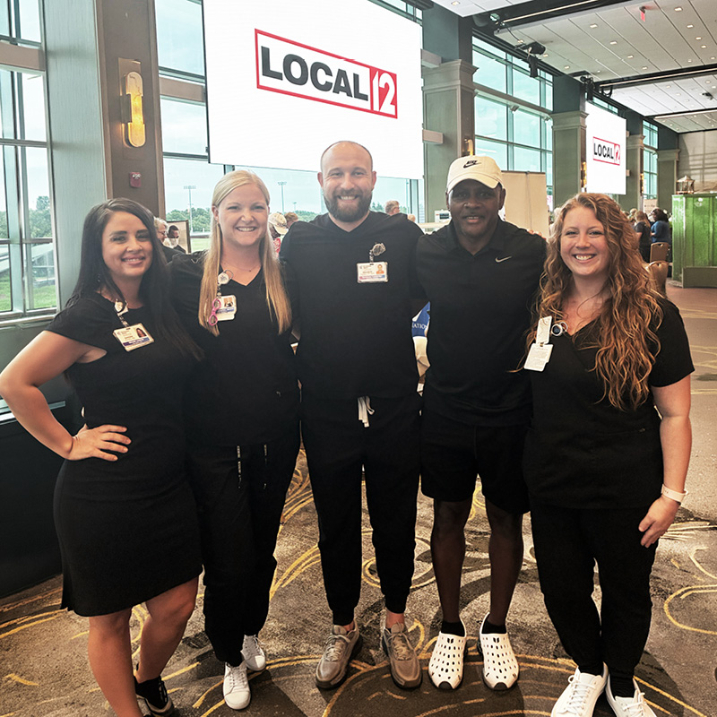 Five healthcare team members pose together at a community event with Local 12 branding displayed on large screens behind them.