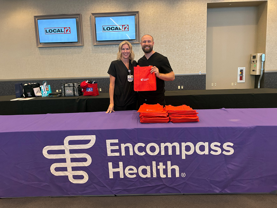 Two representatives staffing an Encompass Health booth display a red tote bag at a community event with Local 12 signage in the background.