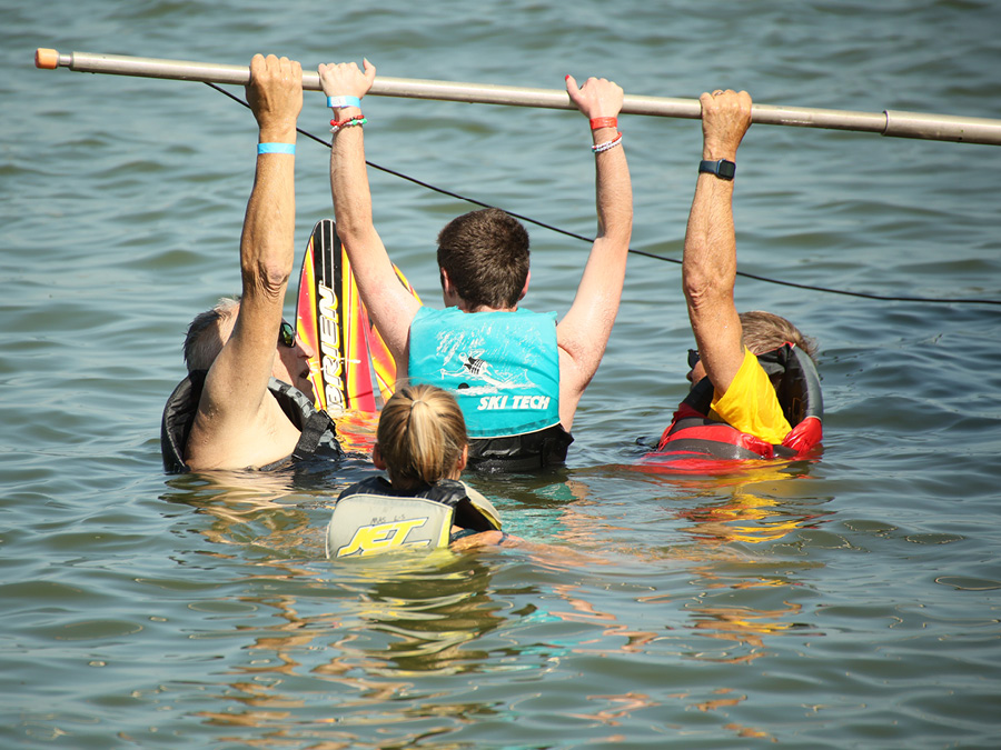Three people wearing life jackets stand in a lake holding onto an overhead training bar, while another person assists nearby. A water ski is partially visible behind the group.
