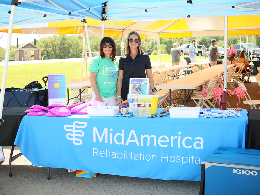 Two people stand behind a MidAmerica Rehabilitation Hospital information table under a canopy at an outdoor event. The table displays promotional materials, giveaways, and bottled water, with seating and attendees visible in the background.