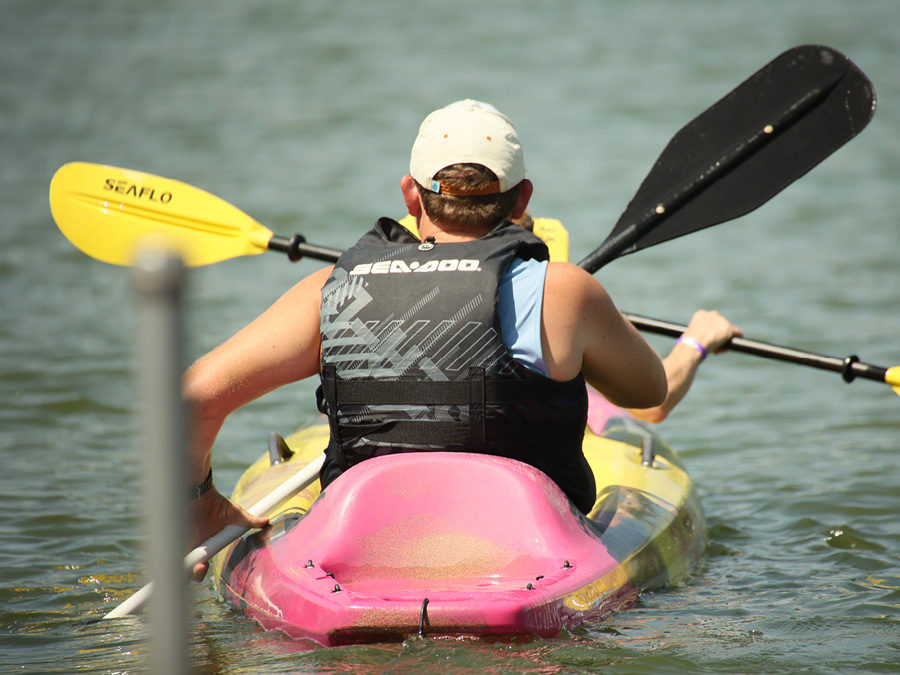 A person wearing a life jacket paddles a colorful kayak across a lake using a double-bladed paddle. The image is taken from behind, with calm water visible in the background.