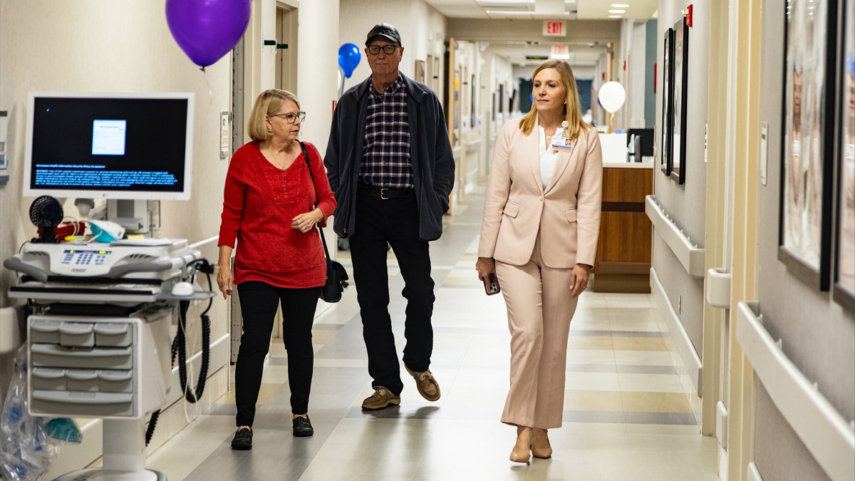 Three people walk through a hospital hallway during a facility tour, passing medical equipment and celebratory balloon displays.