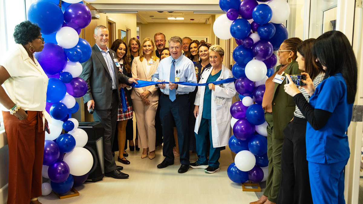 Healthcare leaders and staff participate in a ribbon-cutting ceremony beneath a balloon arch in a hospital corridor.