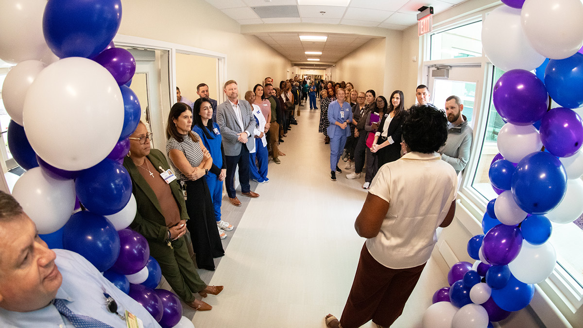 Healthcare staff gather in a hospital hallway beneath balloon arches as a speaker addresses the group during a facility opening event.