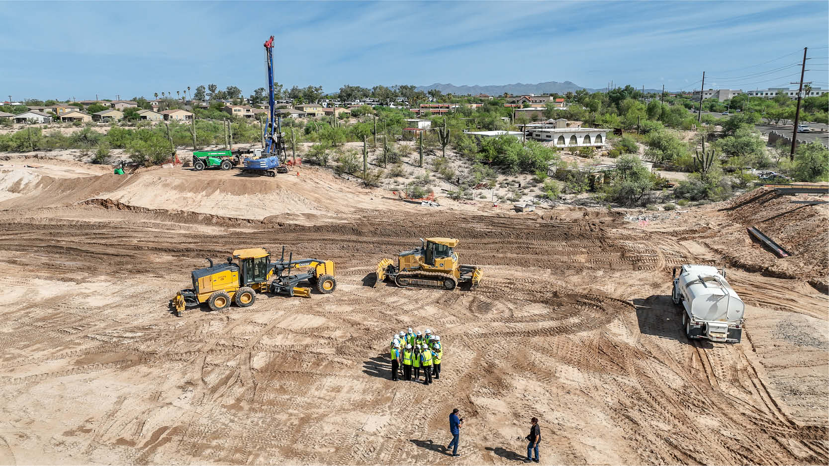 Aerial view of a construction site with heavy equipment and a group of hard-hat-wearing participants gathered during a groundbreaking event.