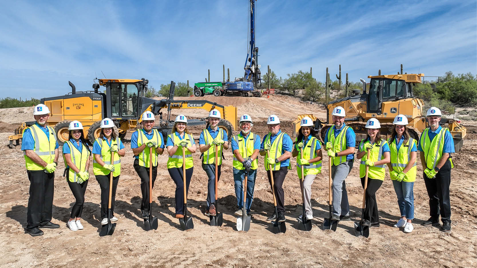 Groundbreaking participants in safety gear pose with ceremonial shovels at a construction site with heavy equipment in the background.