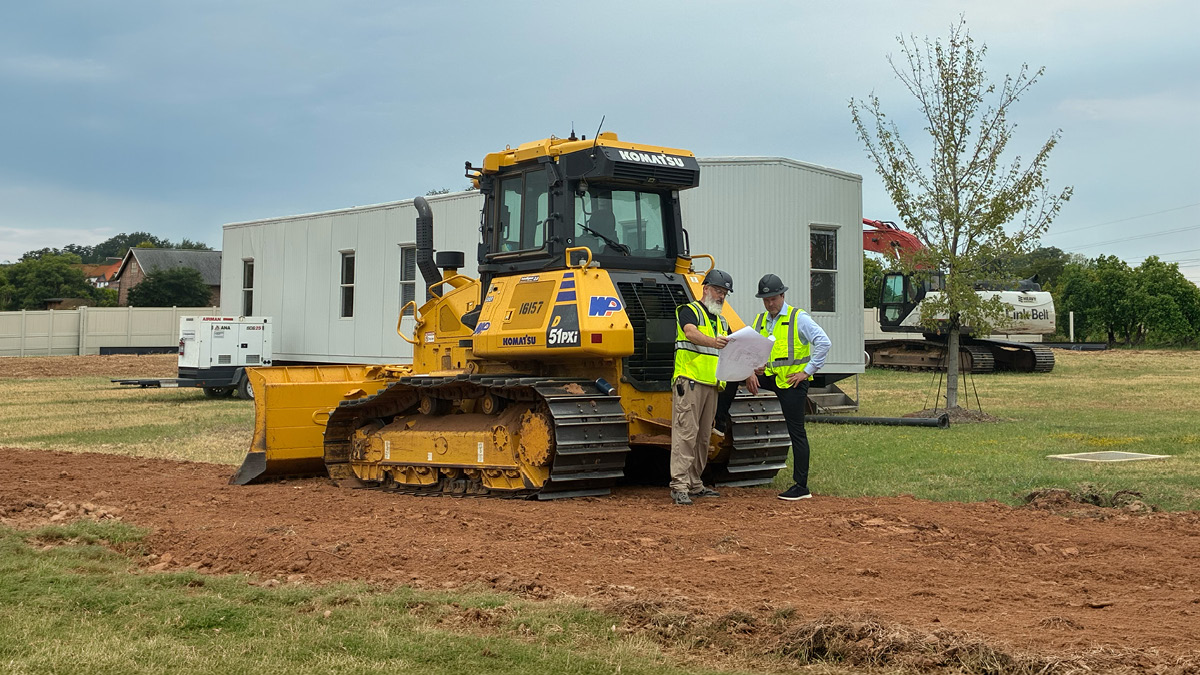 Two construction workers review plans next to a yellow bulldozer at an active construction site.