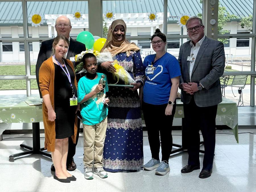 Group photo from a healthcare recognition ceremony featuring Zenab Kone, Daisy Award recipient, holding flowers and an award alongside colleagues and a child holding a trophy.