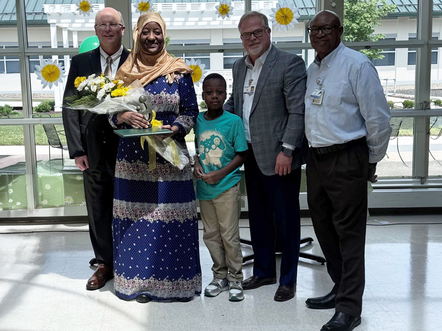Zenab Kone, Daisy Award recipient, poses with flowers and a recognition award alongside colleagues and a child during a healthcare recognition ceremony.