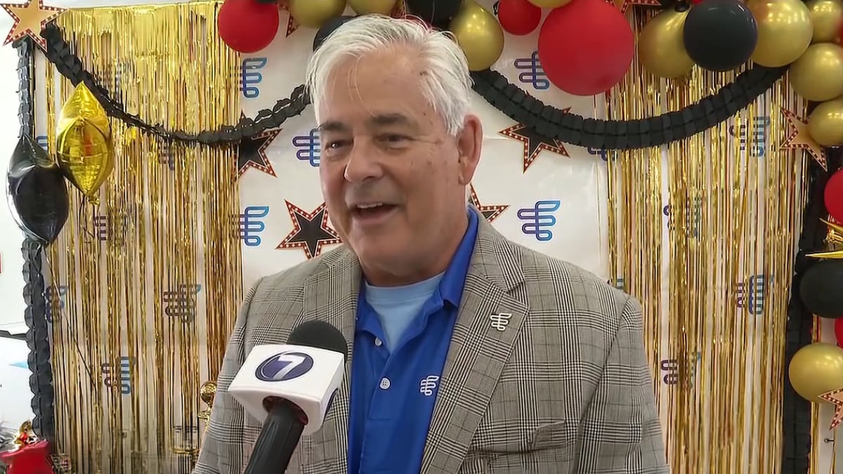 A person is interviewed by a local news station at a themed event decorated with red, black, and gold balloons and gold fringe.
