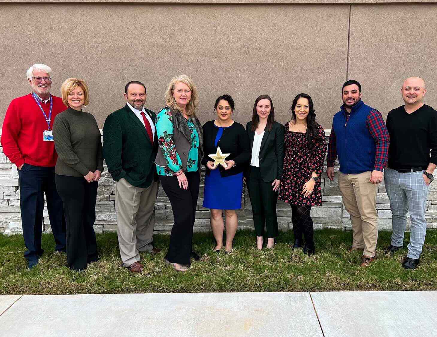 A group of nine people stands outdoors, smiling, in front of a light-colored wall. One person holds a gold star award. The setting conveys a celebratory mood.