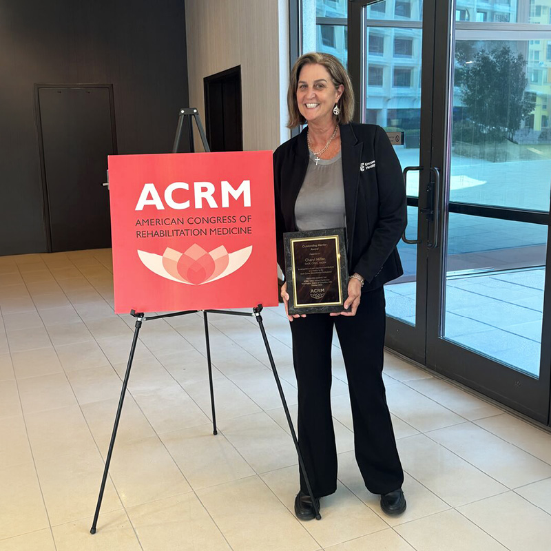 Cheryl Miller poses with an award plaque beside an American Congress of Rehabilitation Medicine (ACRM) sign at a professional event.