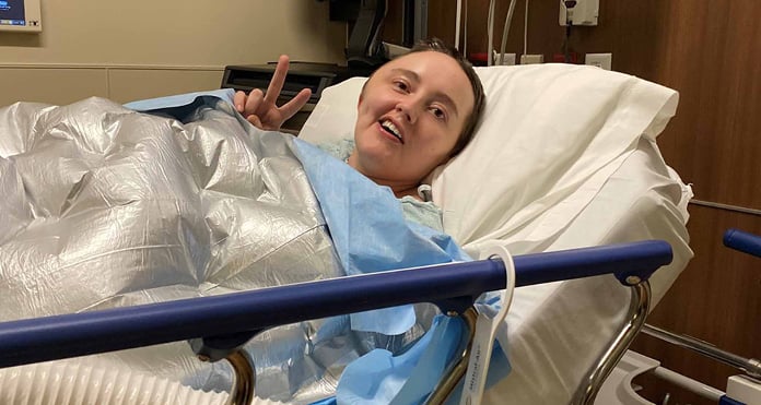 Jessica, recovering from a stroke at 34, rests in a hospital bed covered with a warming blanket and raises her hand in a peace sign.