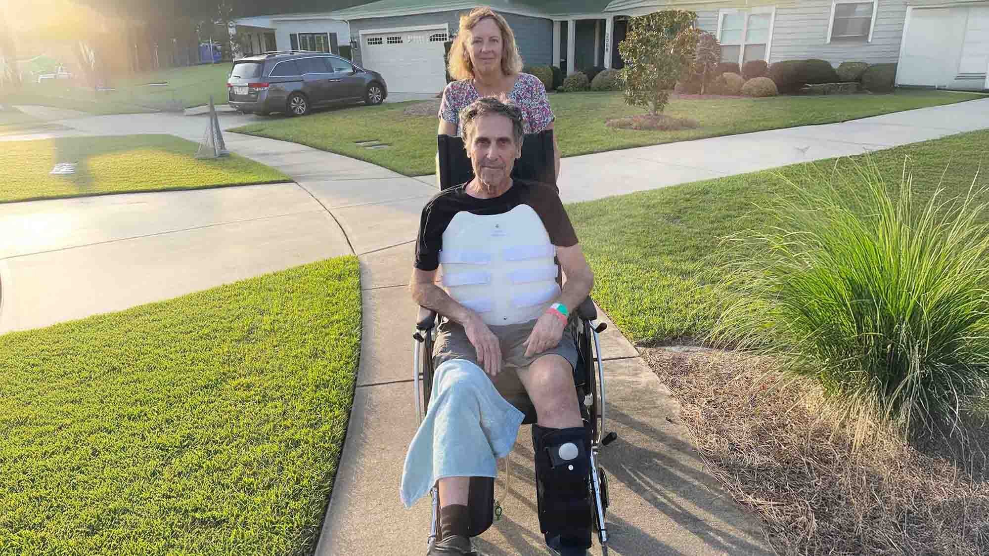 A person sits in a wheelchair wearing a medical brace and leg support while another individual stands behind, pictured outdoors during Kevin’s spinal cord injury recovery.