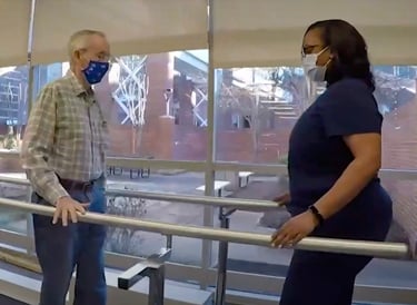 A clinician assists a patient walking between parallel bars in a bright therapy area with large windows, illustrating Martin’s stroke recovery progress.