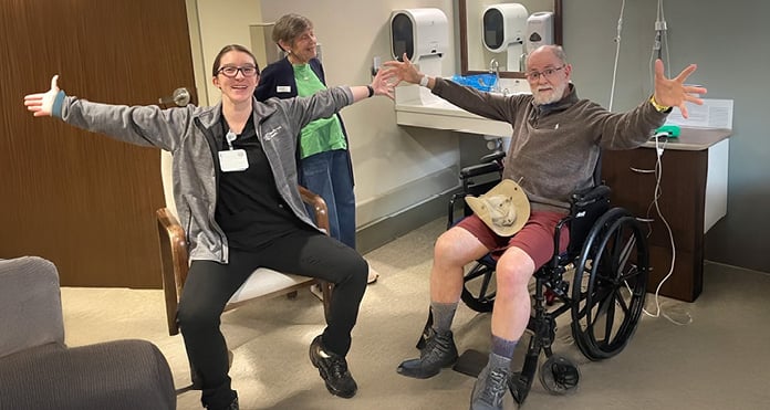 A joyful scene in a medical setting: a smiling woman and an older man in a wheelchair extend their arms wide, expressing happiness and camaraderie.