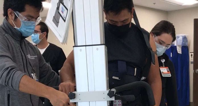 Danilo Sangco is using a walking-assistance device, supported by two healthcare workers in masks. The setting is a clinical environment, conveying a supportive and focused atmosphere.