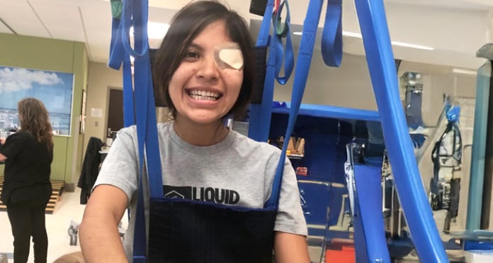 Smiling person with an eye patch, seated in a blue mobility harness, in a rehabilitation setting. The mood is optimistic and encouraging.